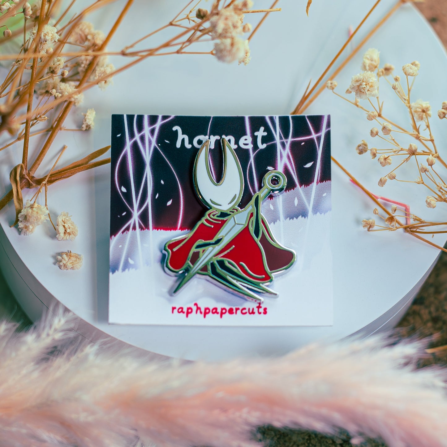 Enamel pin with 'hornet' design on a white surface surrounded by dried flowers