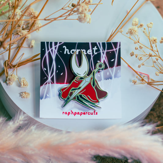 Enamel pin with 'hornet' design on a white surface surrounded by dried flowers