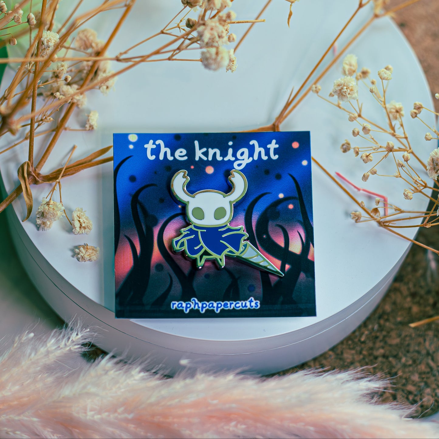 Colorful enamel pin with 'the knight' design on a white surface with dried flowers.