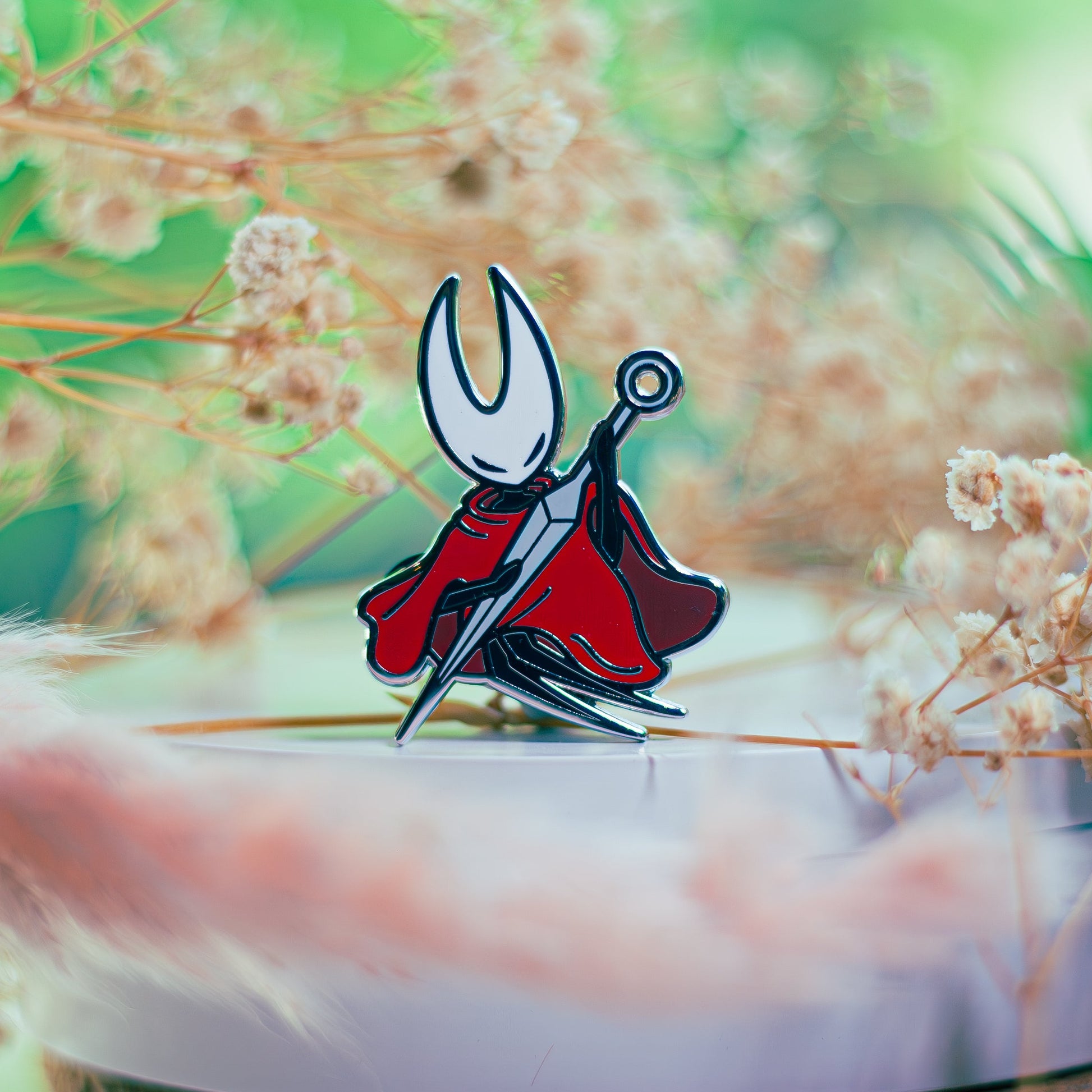 Pin with a design of a character in a red cape and black outfit on a white surface with dried plants in the background.