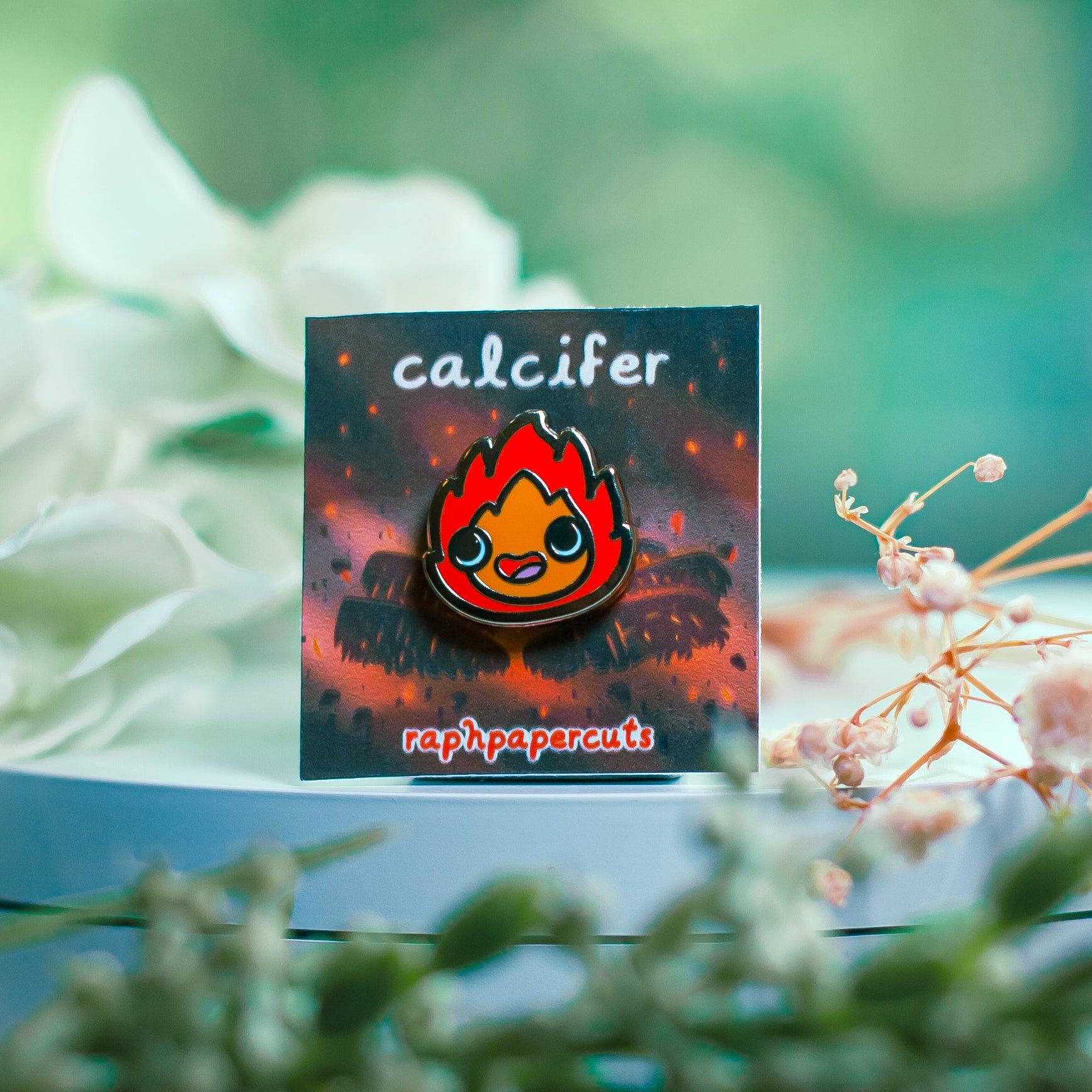 Pin with 'calcifer' design on a card, surrounded by flowers and greenery